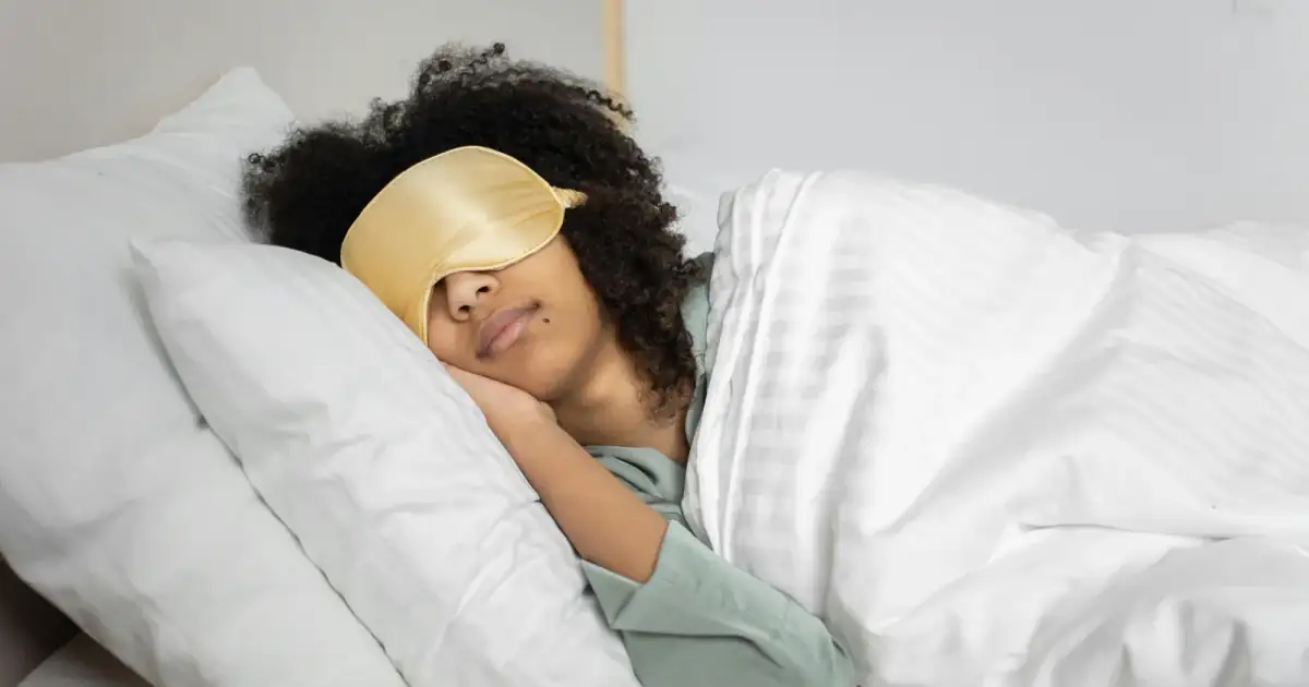 A woman with afro hair sleeps soundly in bed with a sleep mask, enjoying a cozy indoor atmosphere.