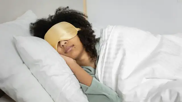 A woman with afro hair sleeps soundly in bed with a sleep mask, enjoying a cozy indoor atmosphere.